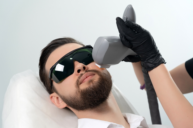 Man lying back wearing protective goggles while a technician uses a laser device on his face for a cosmetic treatment.