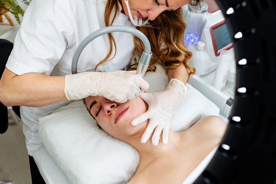 Dental or skincare professional wearing gloves performs a facial treatment on a patient, using a curved handheld device around the mouth.