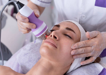 Picture7 | Dr Swetha Cosmodermcentre Esthetician wearing gloves uses a purple handheld facial device on a woman's face during a spa treatment.